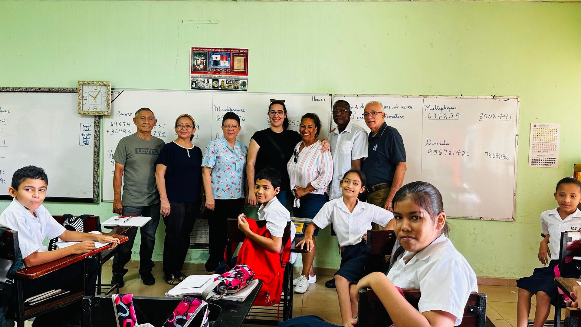 Group of adults and children in a classroom; some smile at the camera, whiteboard and desks visible.