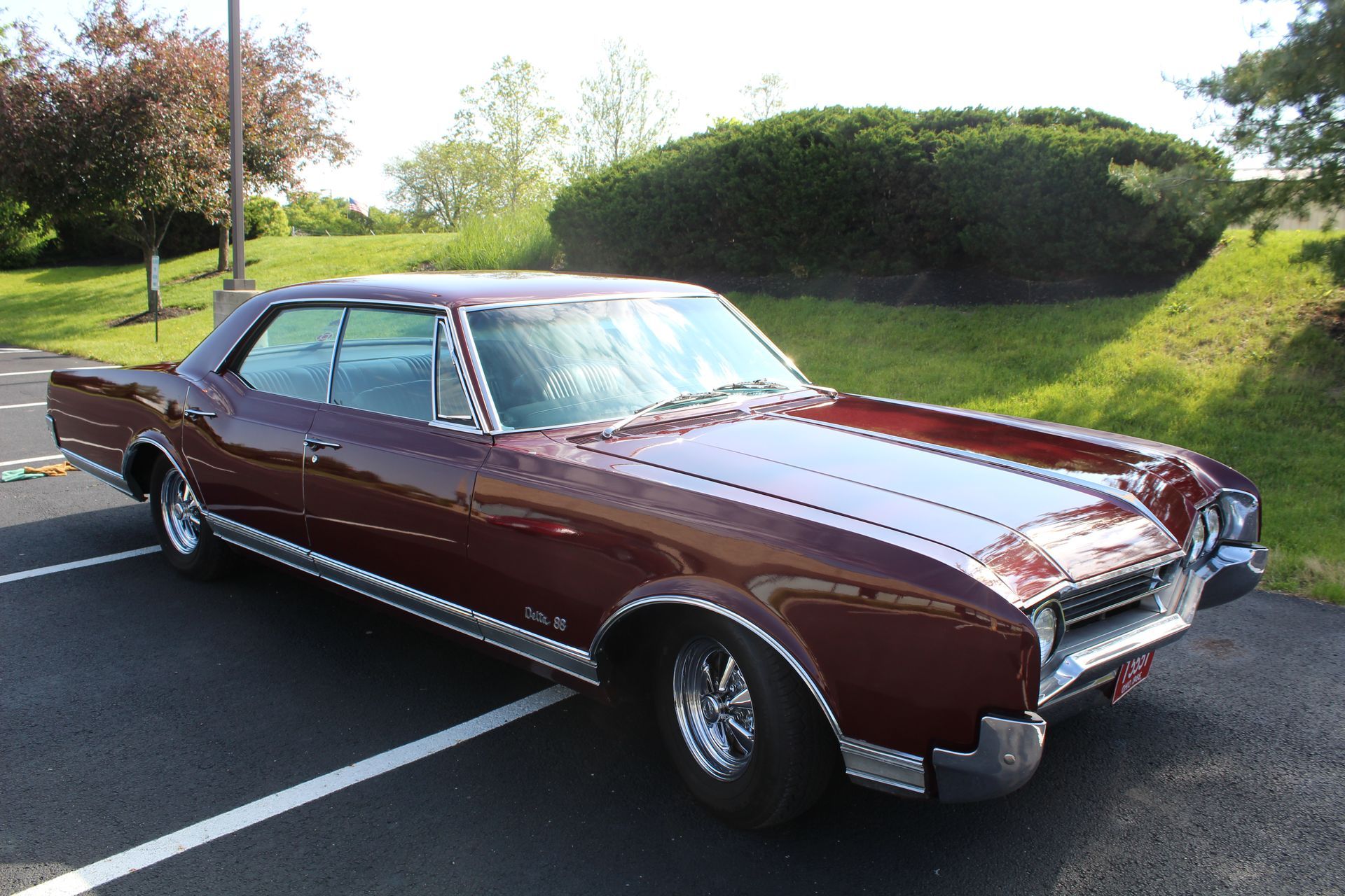 Maroon classic car parked in a lot beside a grassy hill on a sunny day