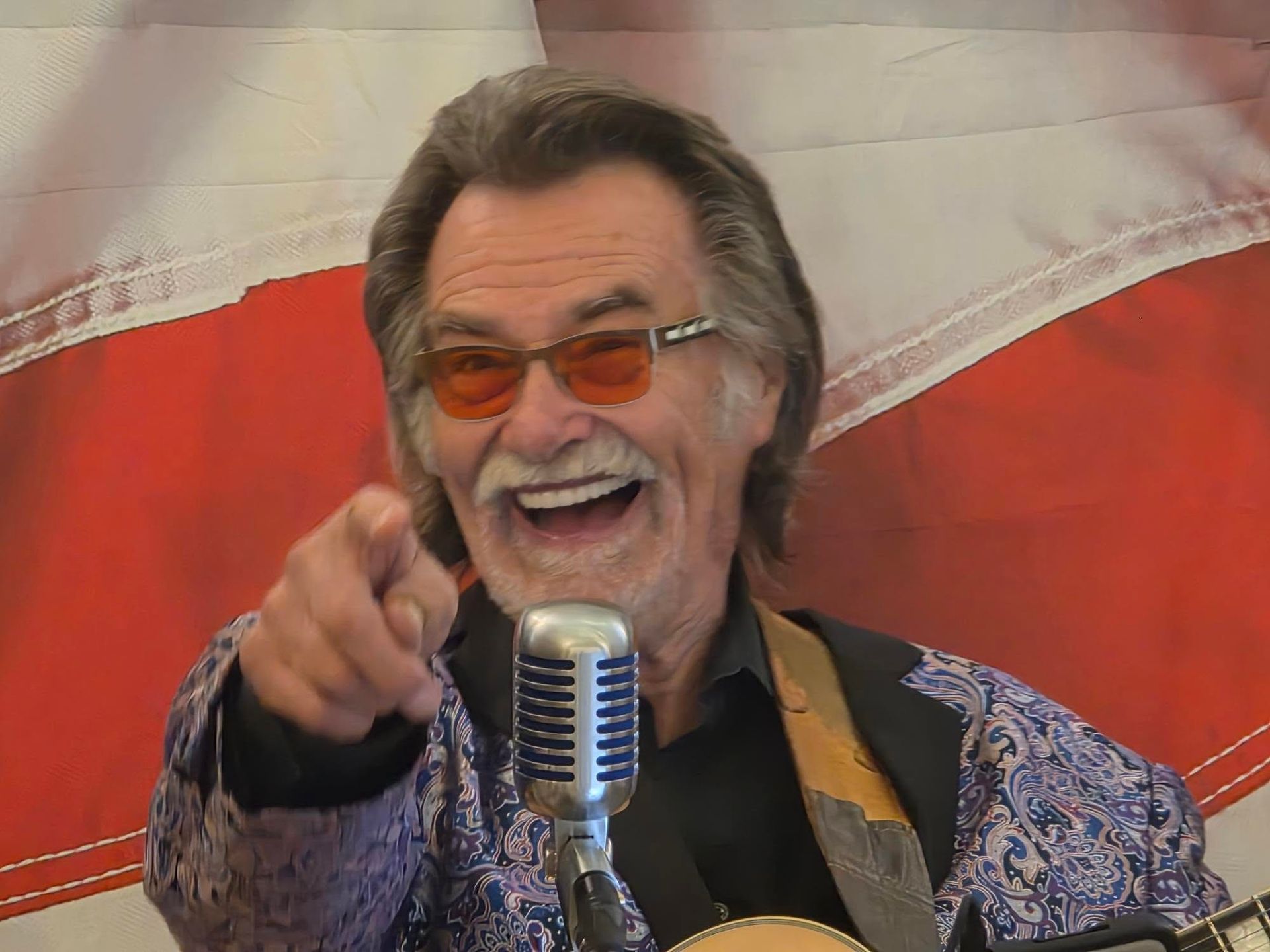 Smiling performer in orange sunglasses points at the camera beside a microphone, with a red-and-white backdrop