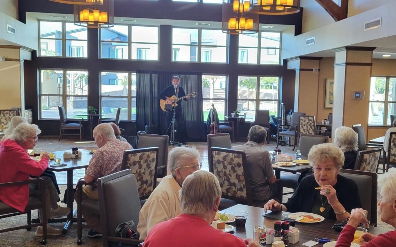 People dining in a bright restaurant while a musician performs on a small stage.