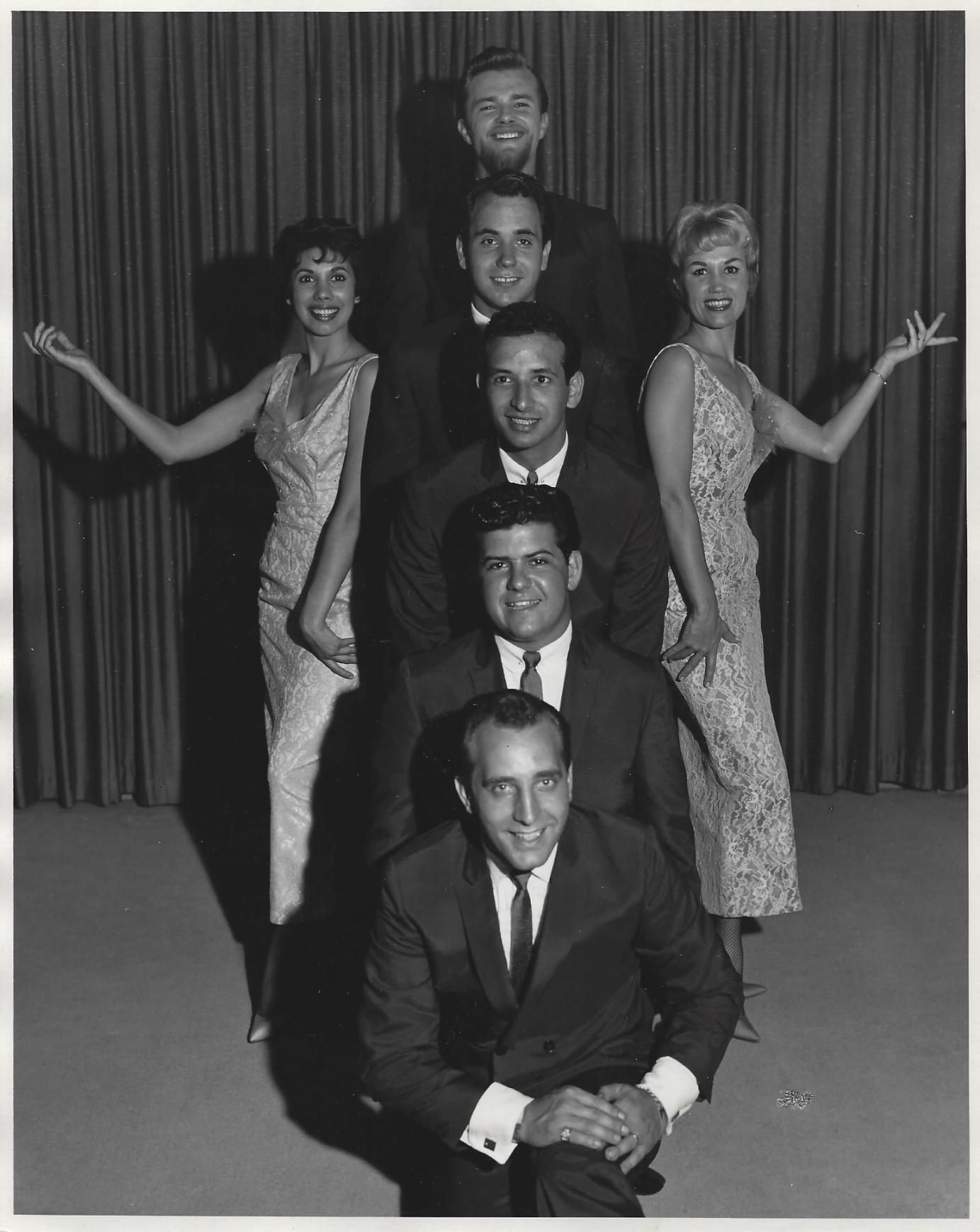Black-and-white portrait of six performers stacked in a vertical pose against a curtain backdrop