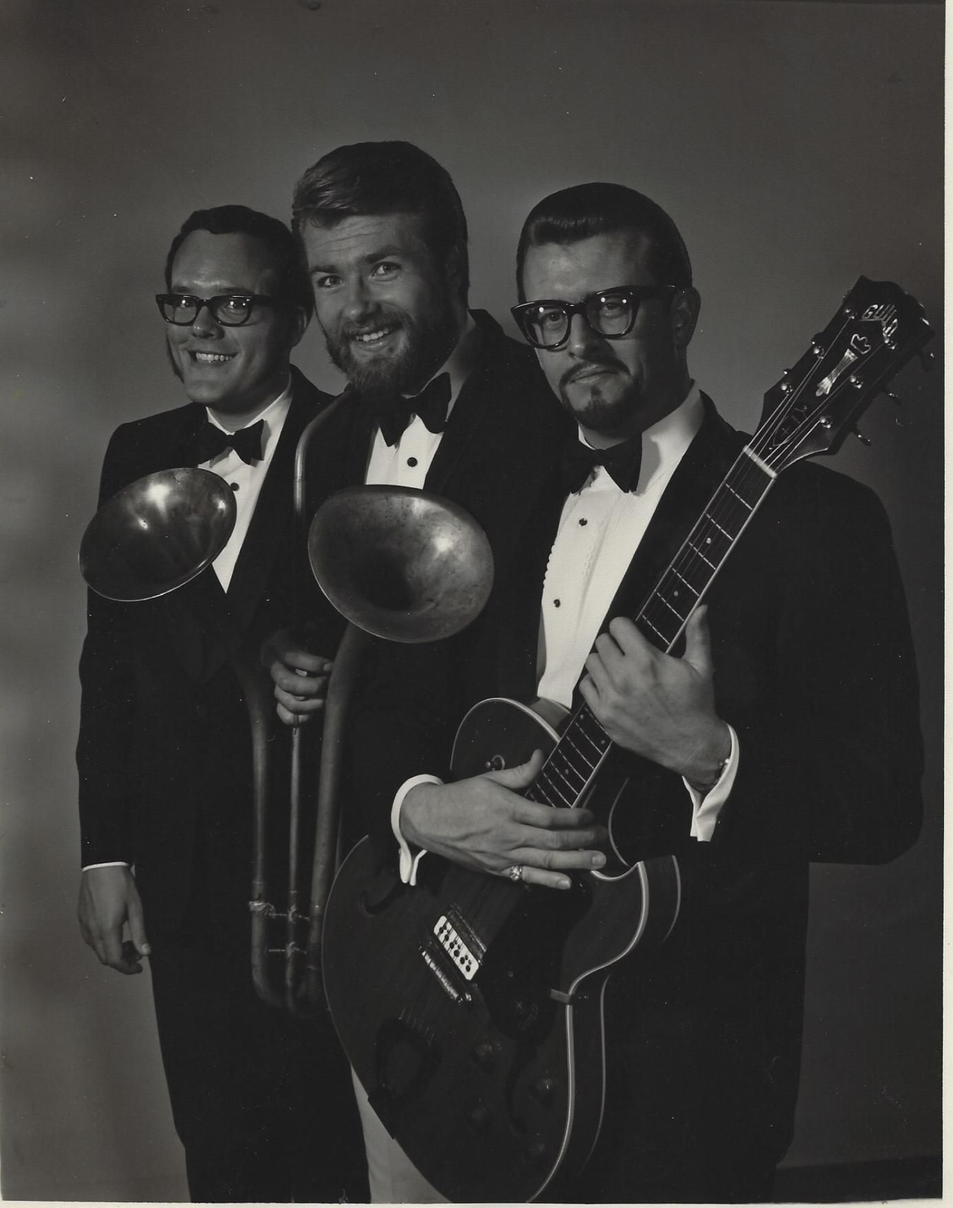 Three men in tuxedos holding trombone, French horn, and guitar in a studio portrait