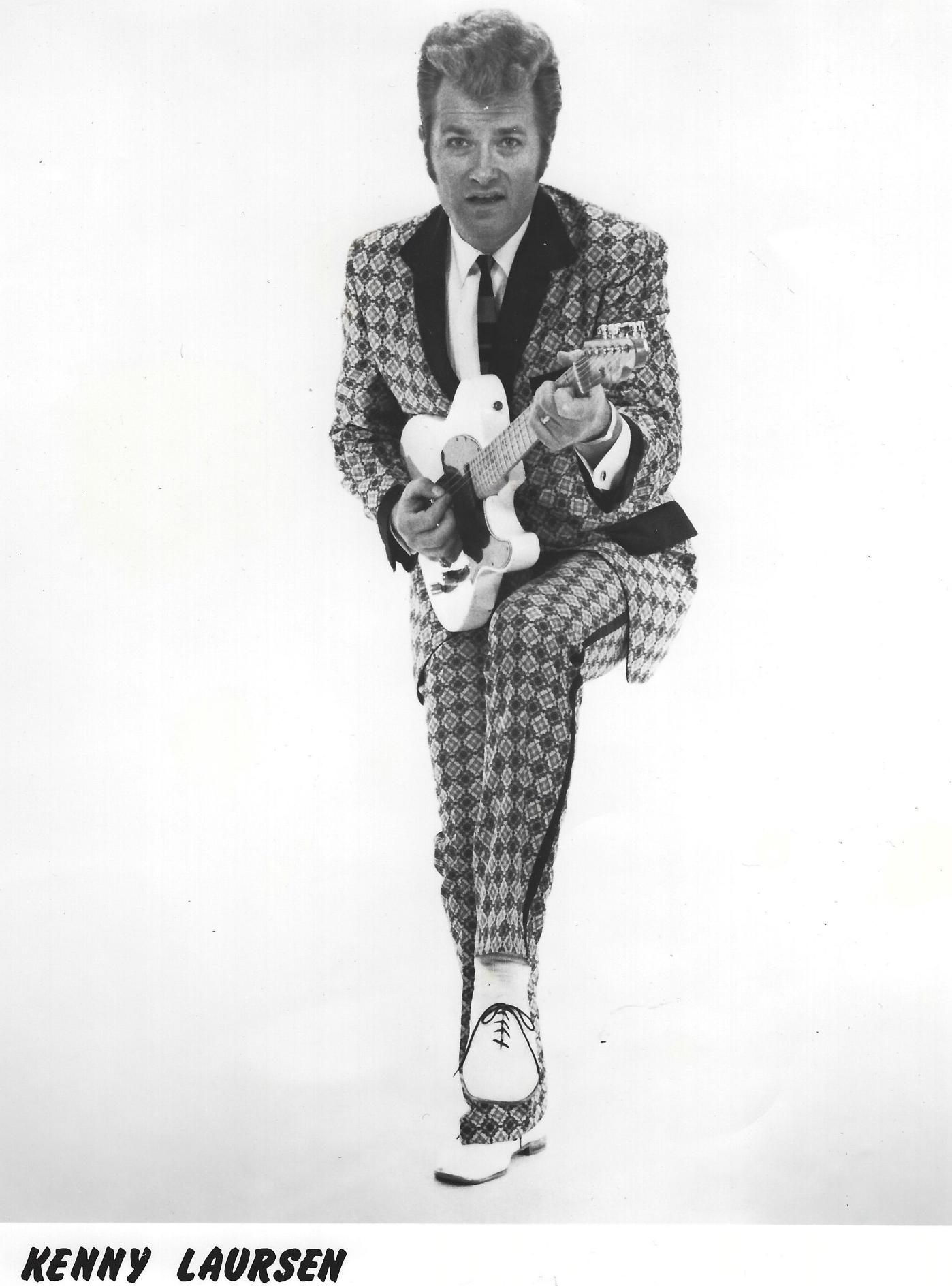 Black-and-white photo of a man in a checkered suit holding a guitar, posed mid-step against a white background