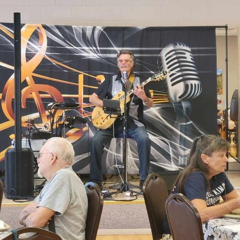 Kenny Laursen playing guitar on a small stage with a large microphone backdrop