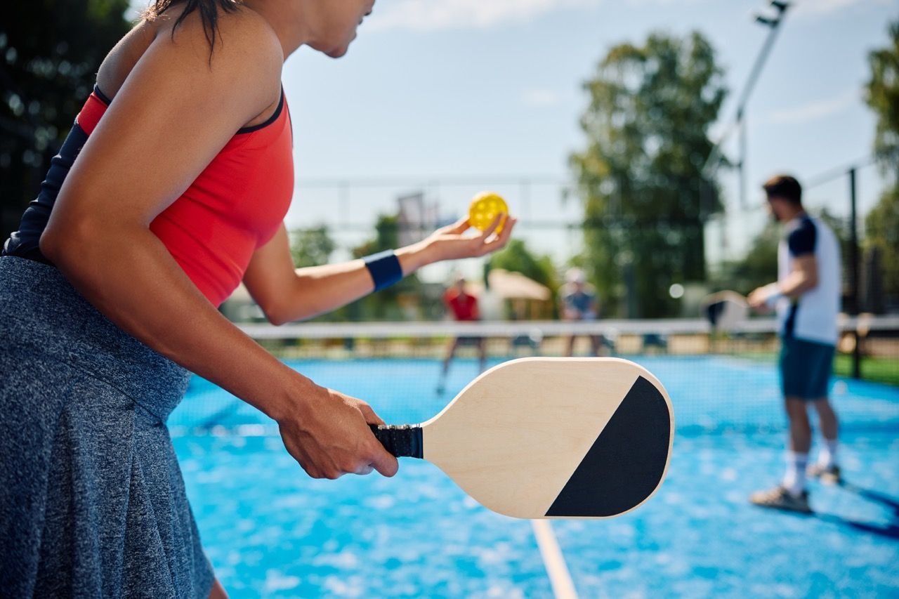 Person holding a pickleball paddle and ball by a pool, with another player in the background.
