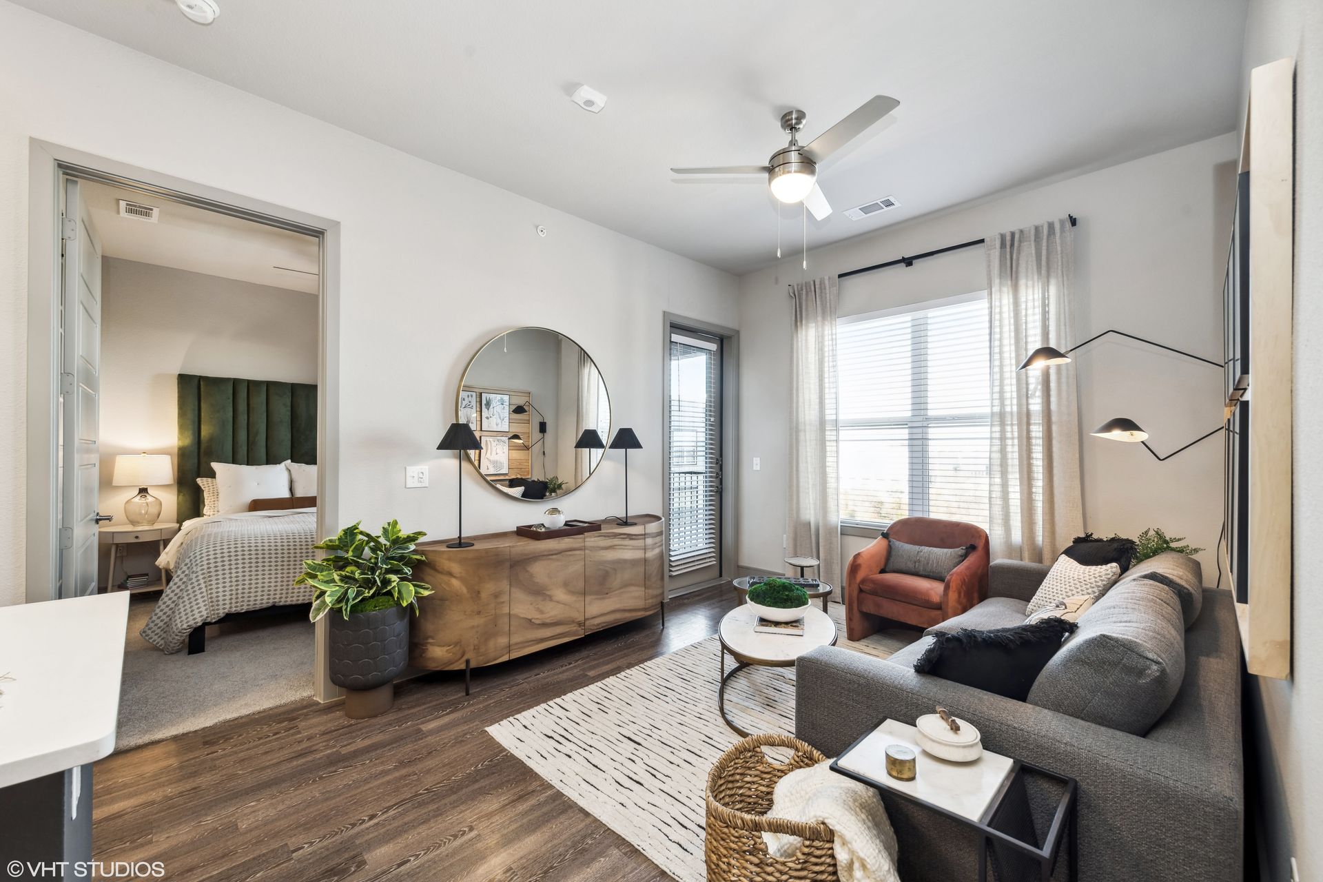 Bright living room with gray sofa, wood dresser, round mirror, armchair, and large window with white curtains