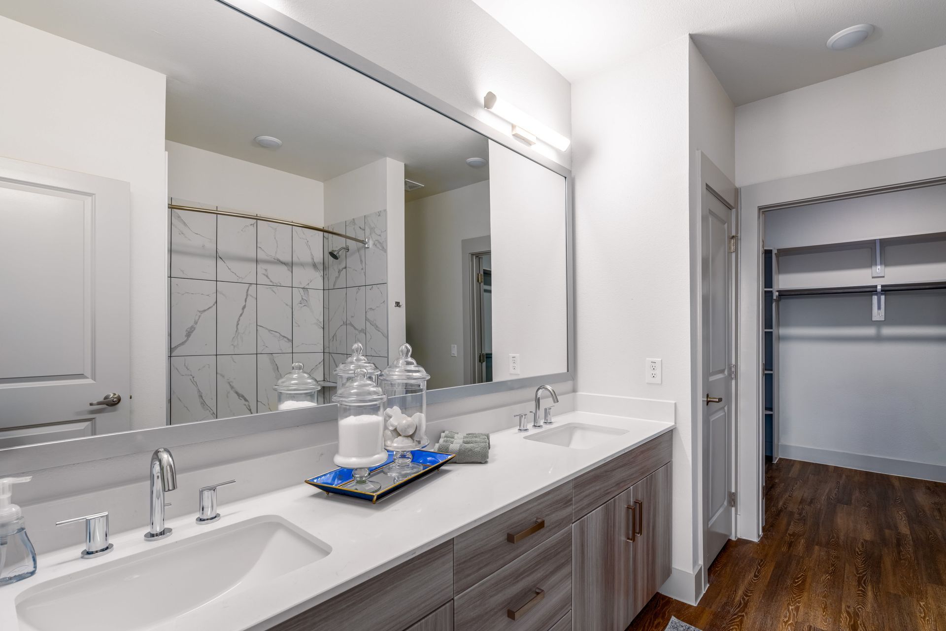 Modern bathroom with double vanity, large mirror, and open closet with hardwood floors