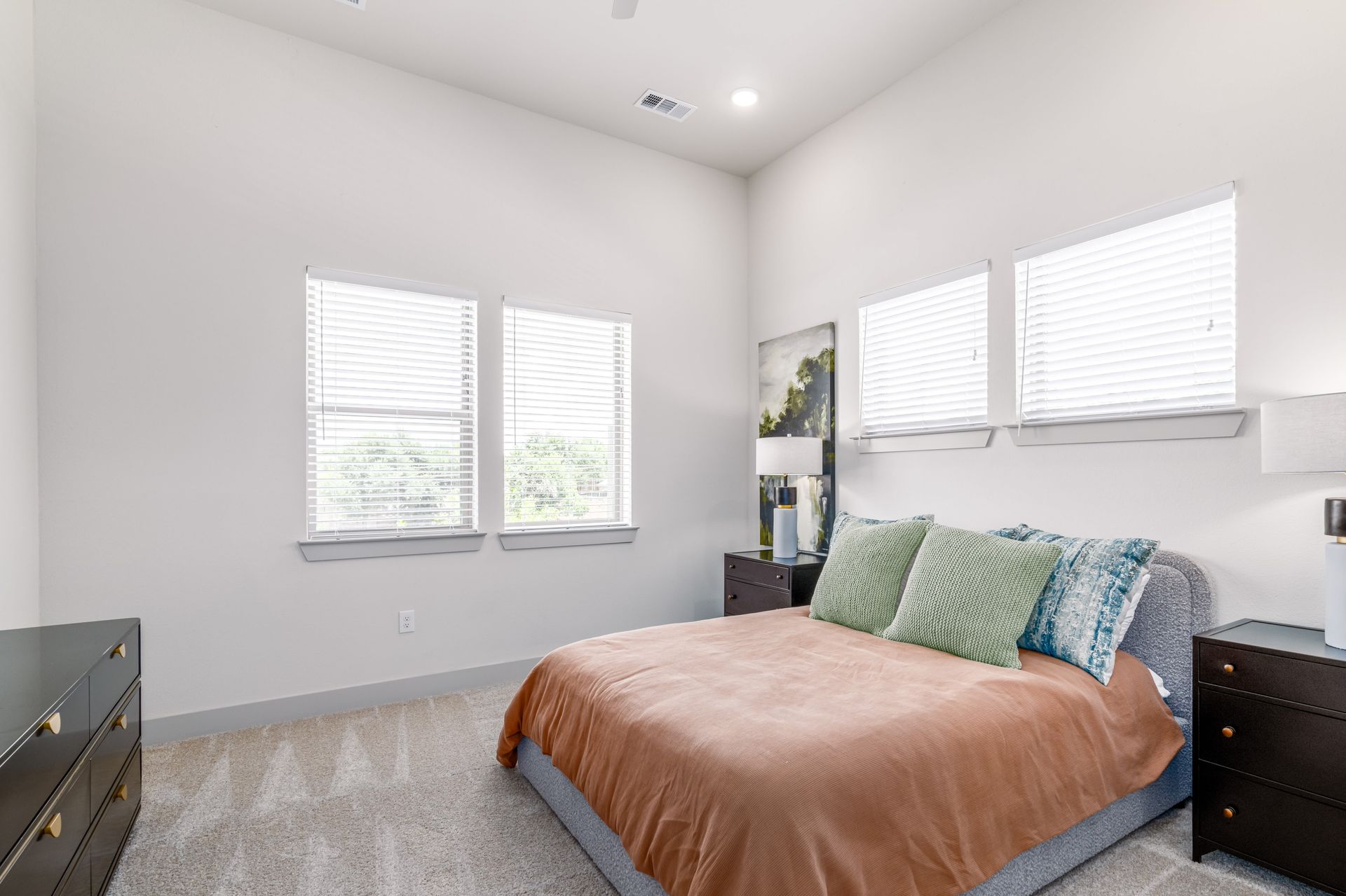 Bright bedroom with a bed, white walls, gray carpet, and multiple windows with blinds