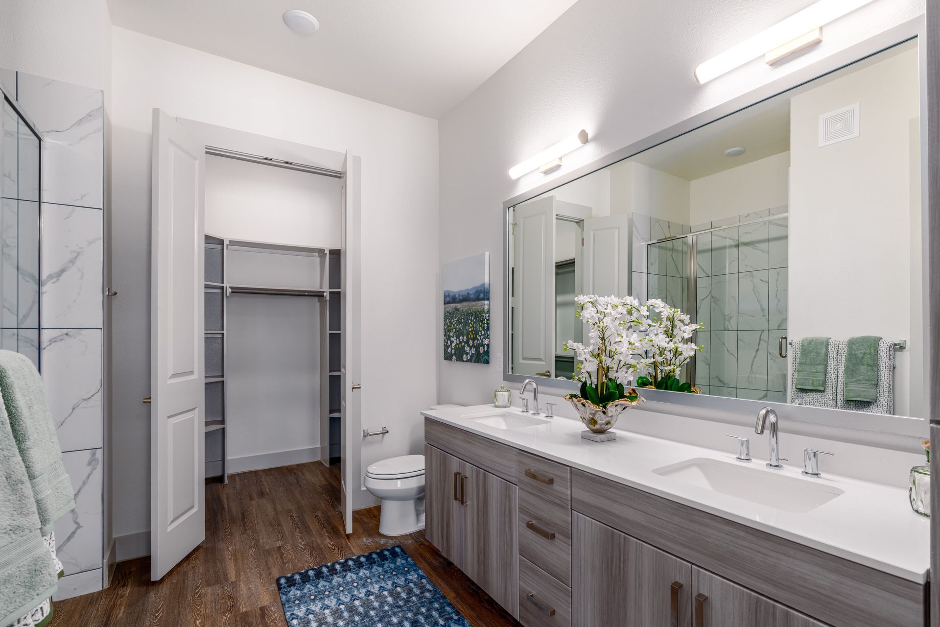 Bright bathroom with double vanity, large mirror, blue rug, and open closet doorway