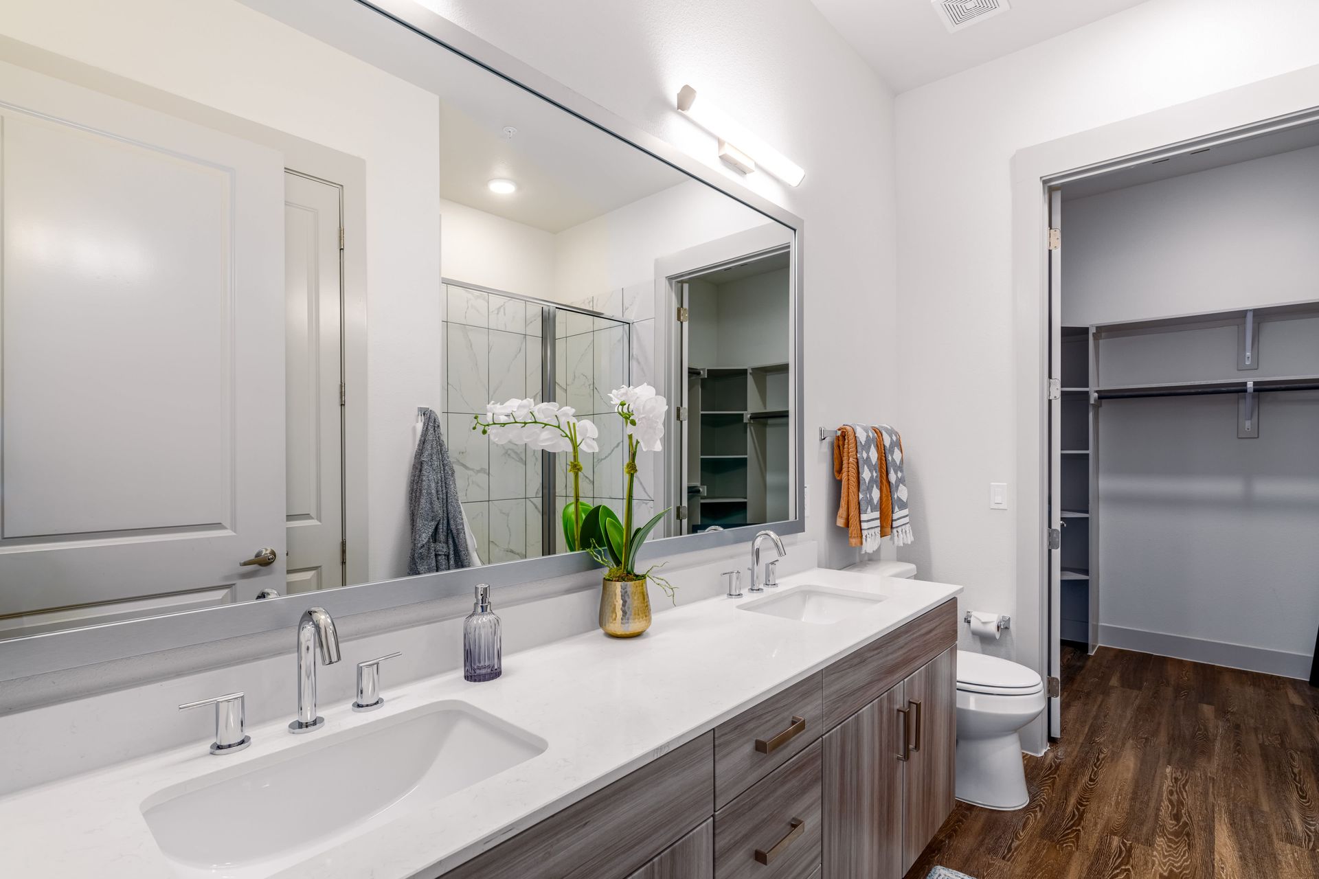 Bright modern bathroom vanity with mirror, double sinks, and an open closet with shelving.