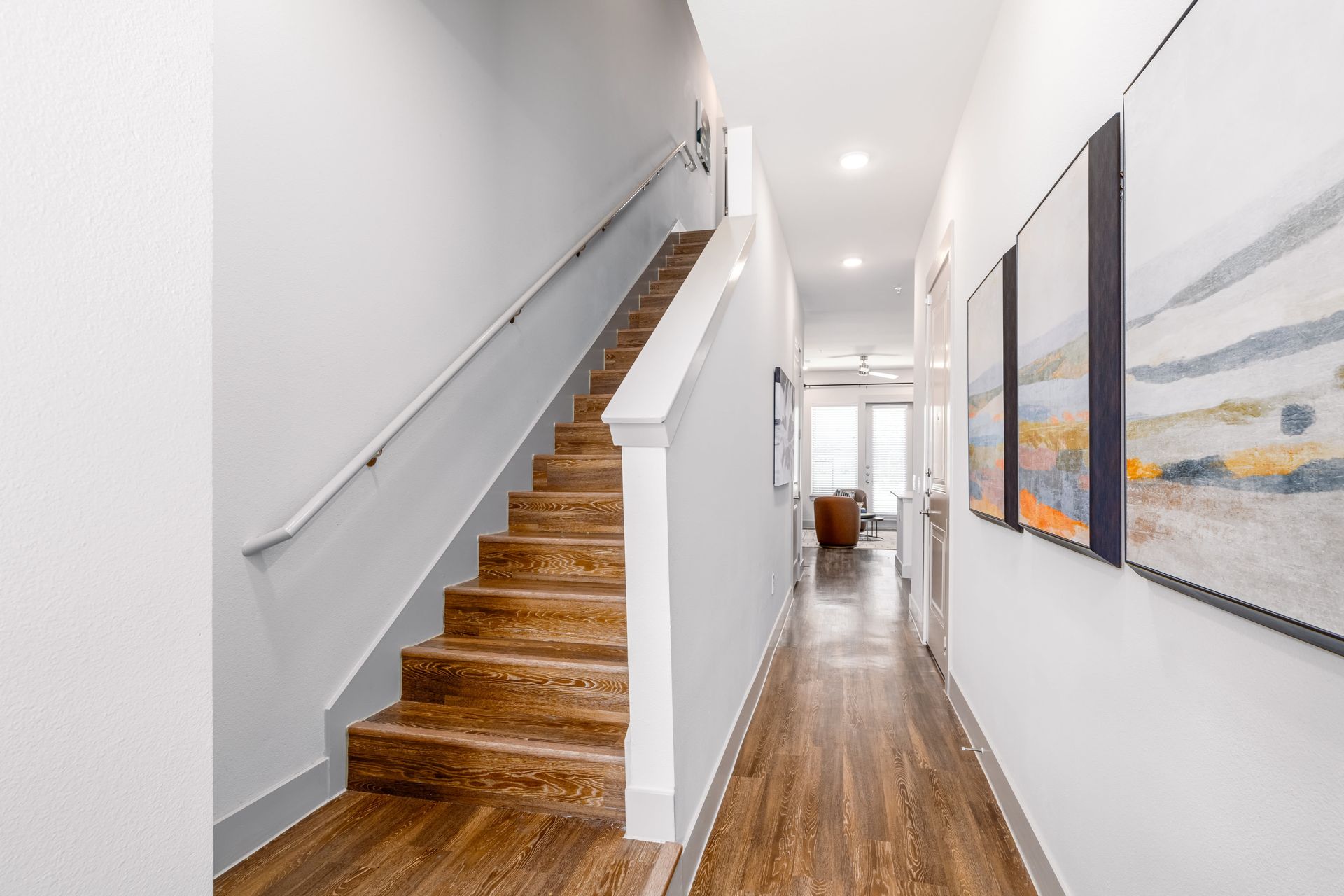 Modern hallway with wooden stairs, white walls, and framed artwork leading to a bright living area.