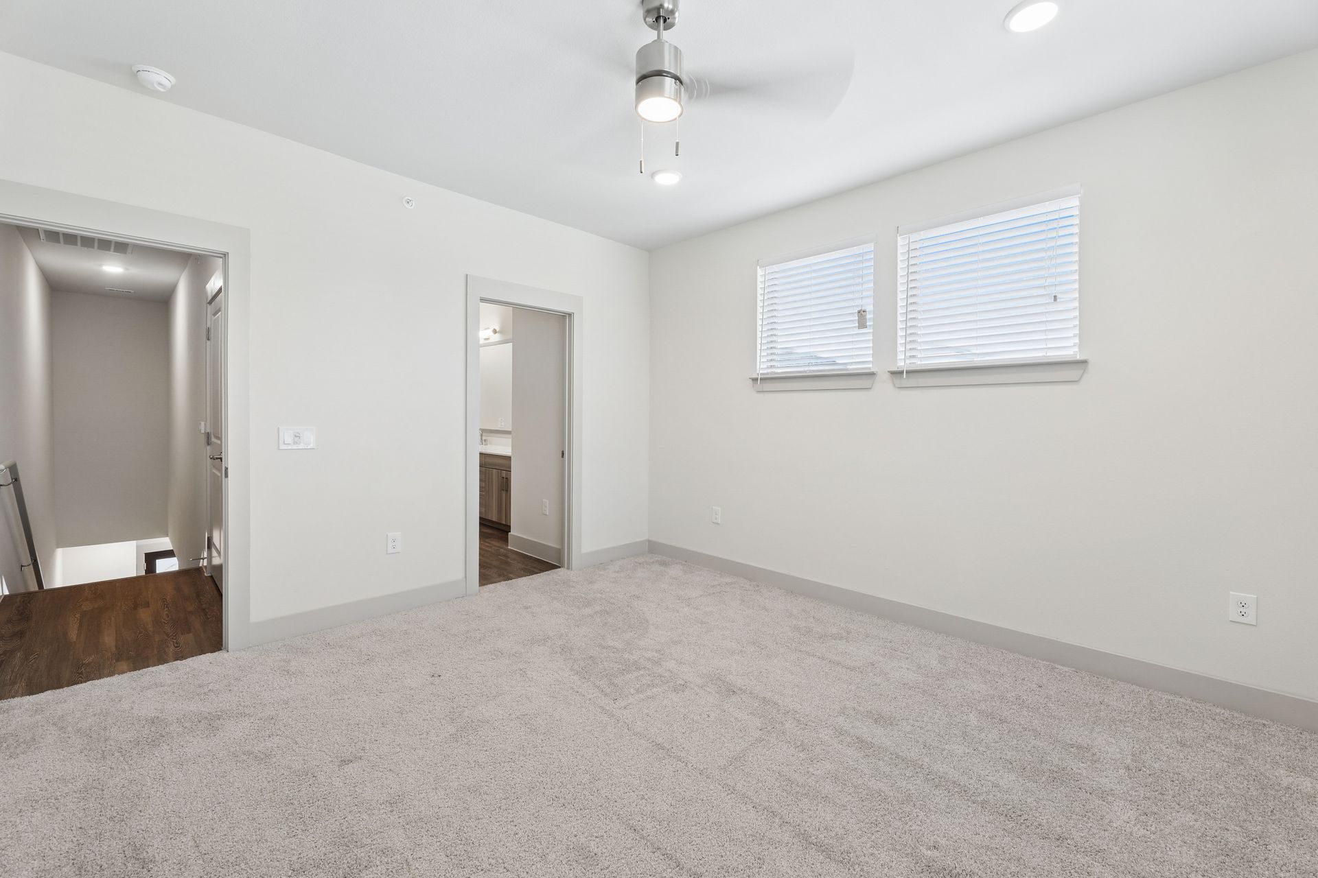 Empty gray-carpeted bedroom with white walls, ceiling fan, two doorways, and a small window