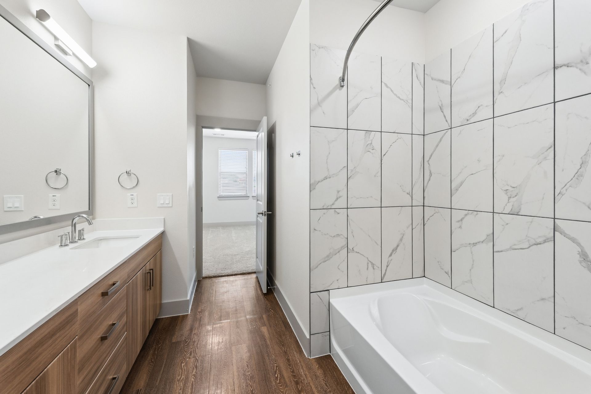 Bright bathroom with wood vanity, large mirror, and marble-look shower tile with tub.