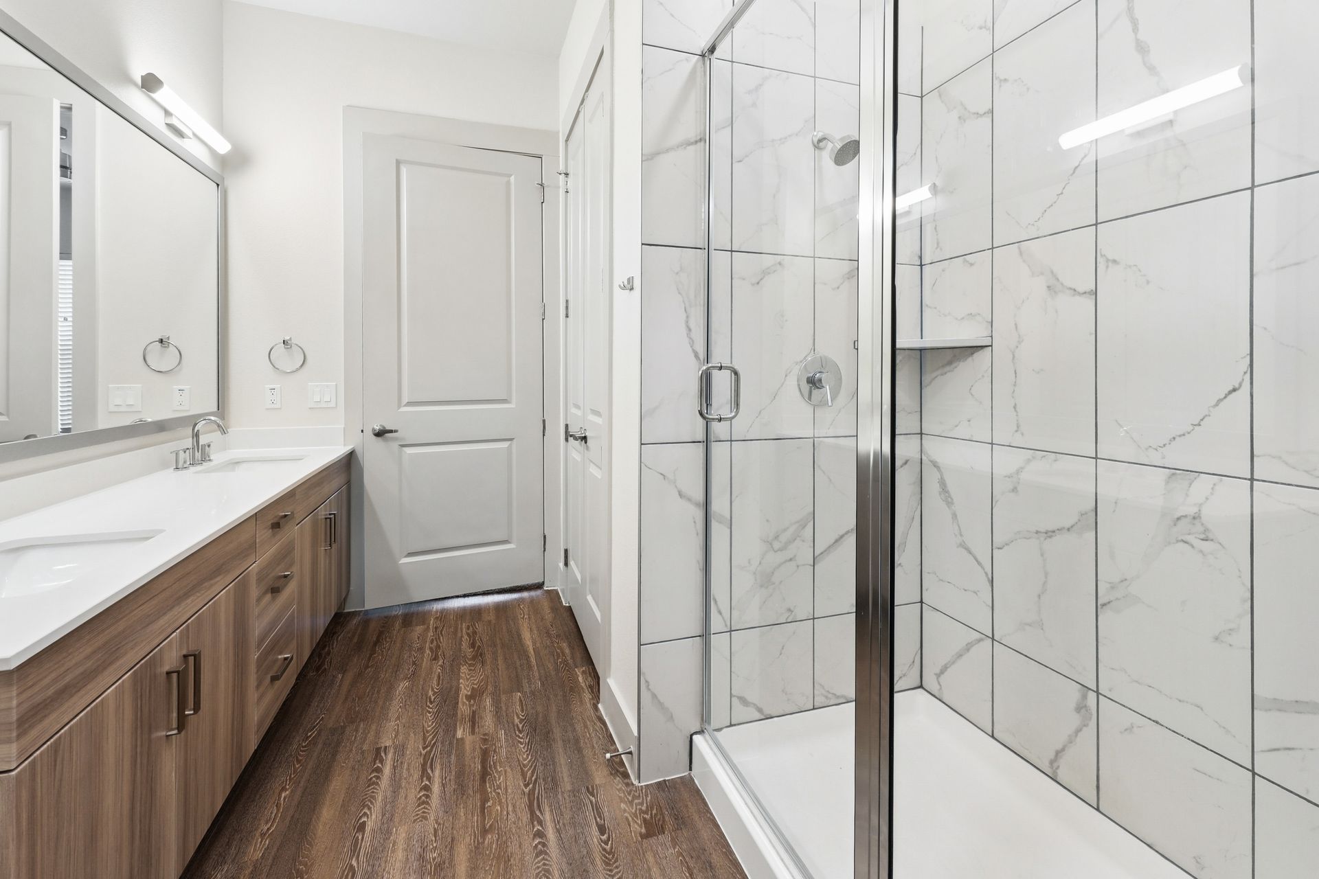 Modern bathroom with double vanity, white marble tile, and glass-enclosed shower with wood-look flooring