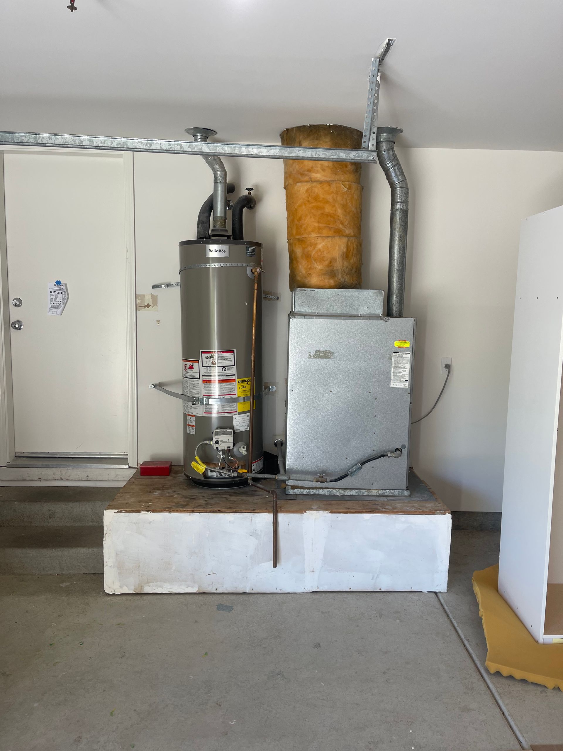 Furnace and water heater on a raised platform in a garage, with ductwork and a door visible.