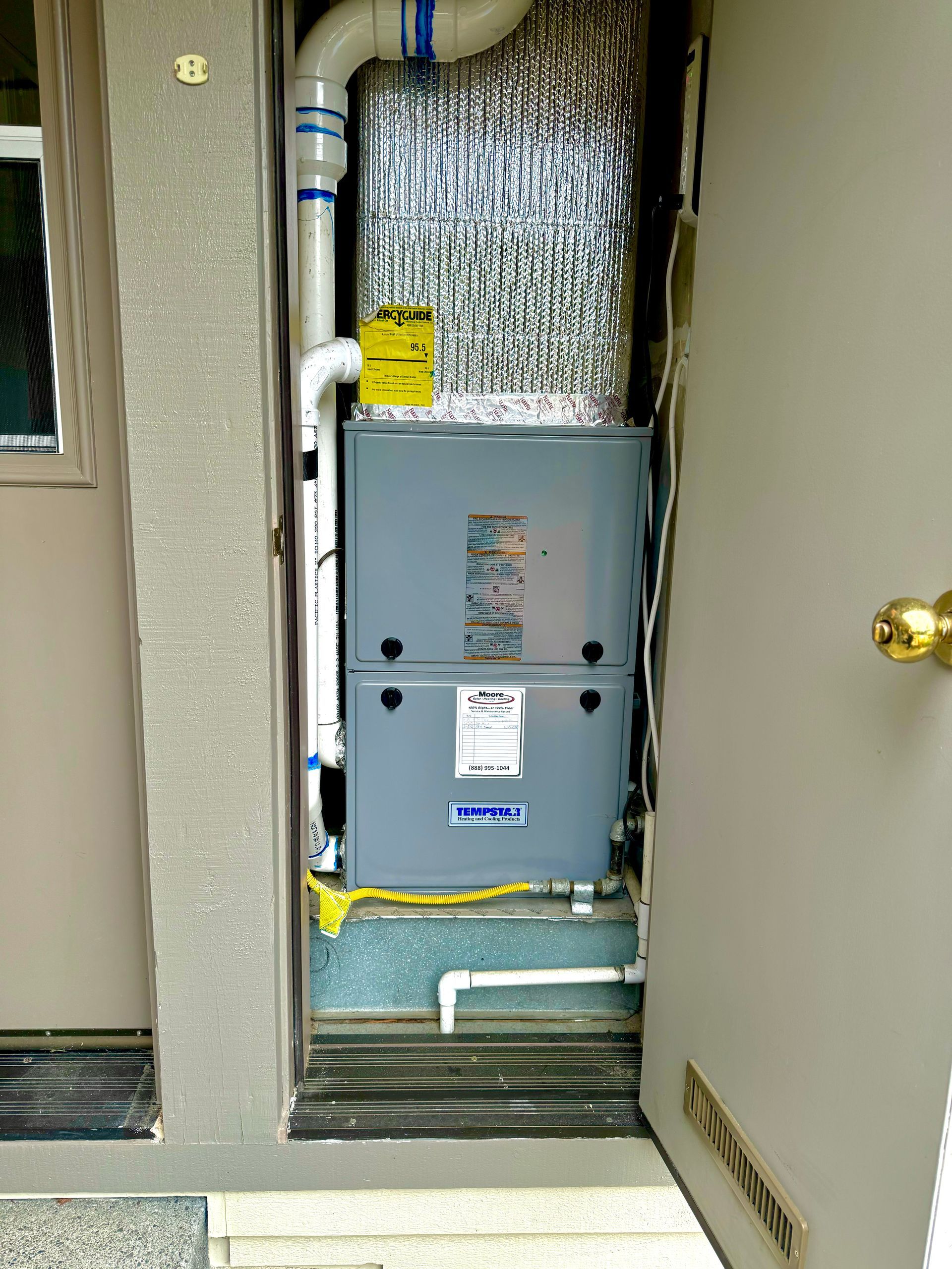 Furnace unit installed in a utility closet with white pipes, silver ductwork, and open door.