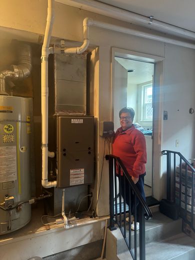 Woman stands near a furnace and water heater in a utility room; doorway and stairs are nearby.