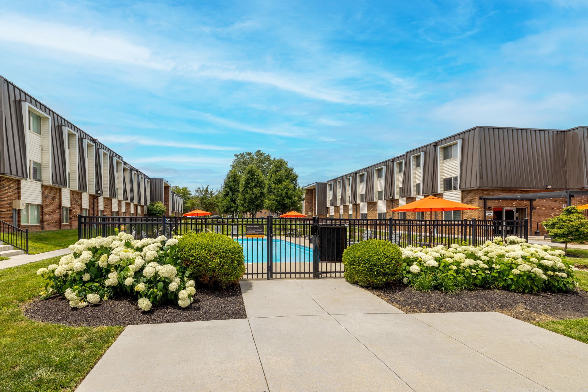 apartment courtyard with pool, flower beds, and orange umbrellas under a blue sky