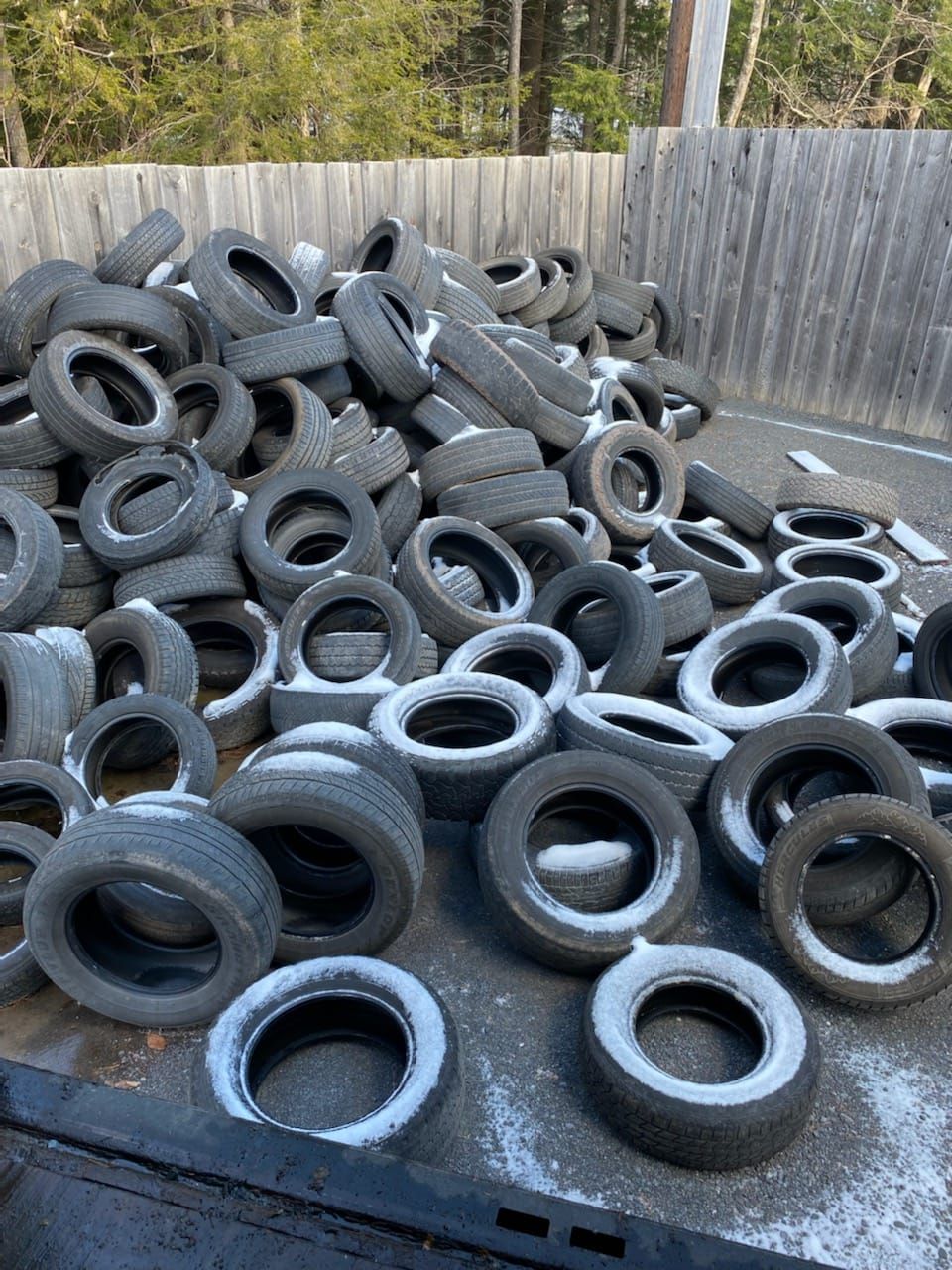Pile of used tires, some with traces of snow, outdoors against a wooden fence.
