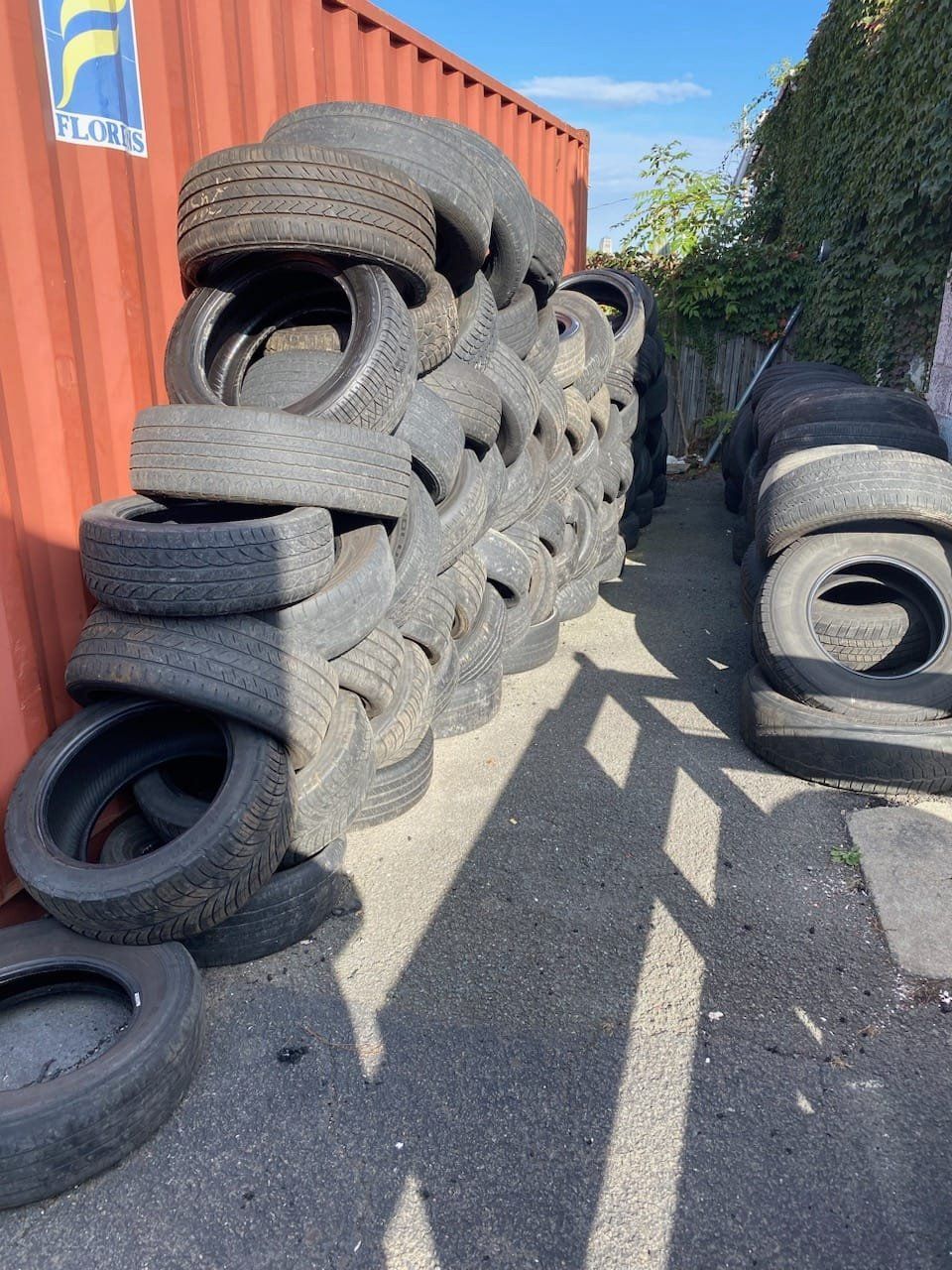 Pile of used tires stacked next to a red container and fence. Asphalt ground.