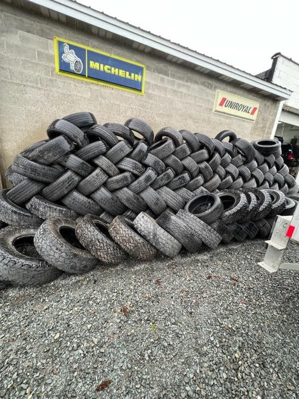 Pile of used tires stacked outside a tire shop, next to a brick wall with a Michelin sign.