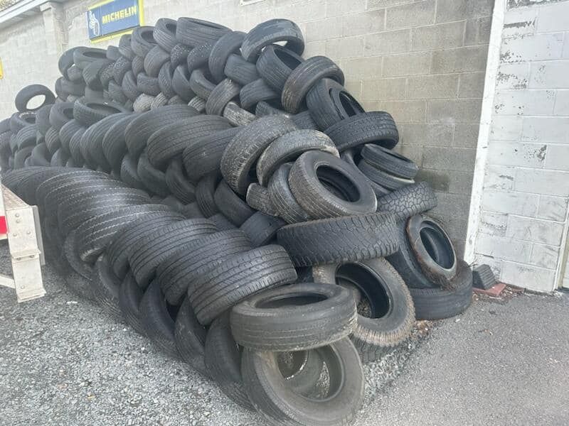 Pile of used black tires stacked against a gray wall in an outdoor setting.