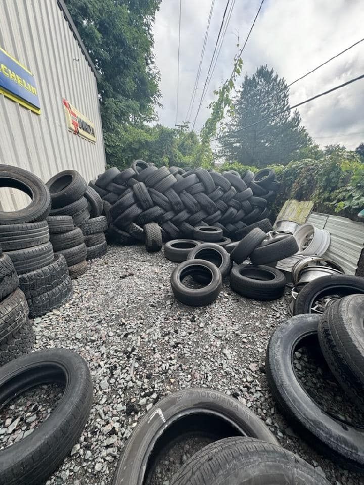 Tires stacked in an outdoor lot, a large pile in the background, cloudy sky.