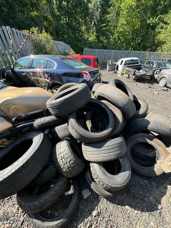 Pile of used tires in junkyard; various cars visible in background. Sunny day.