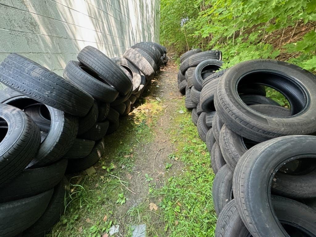 Stacks of used tires line a dirt path between a concrete wall and foliage.