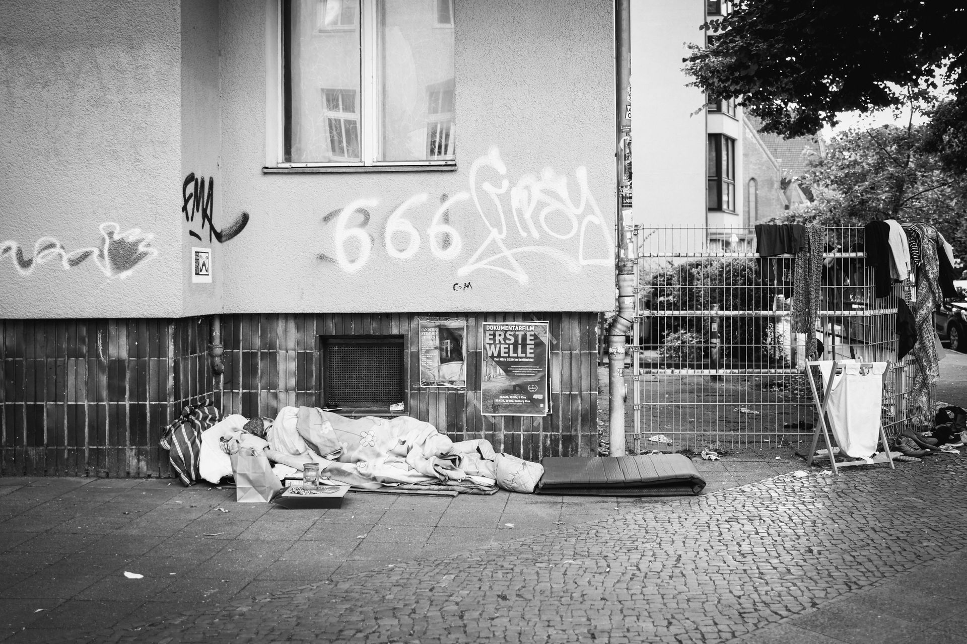 Black and white photo of a building's exterior with graffiti. A pile of belongings and bedding rests on the sidewalk.