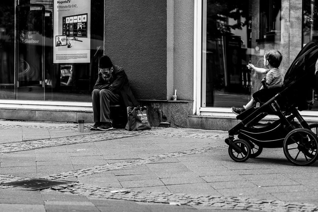 A person sits slumped on a sidewalk, near a baby in a stroller pointing towards a store window.