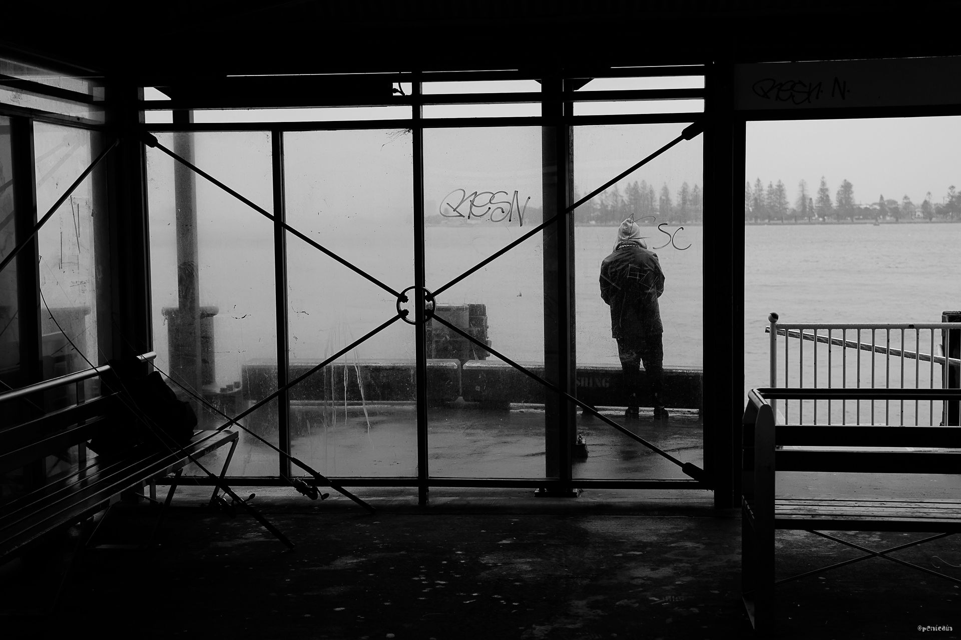 Person in a raincoat on a pier looking out at water through a rain-streaked window.