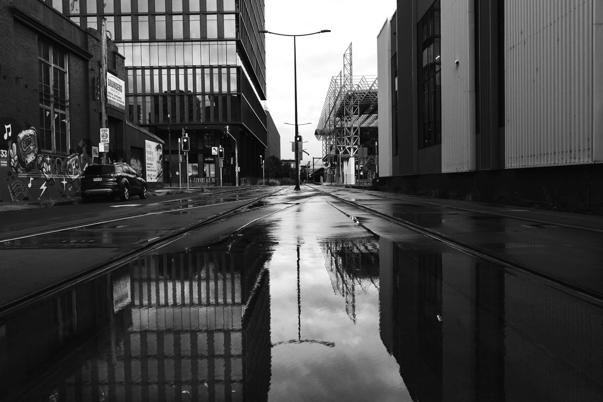 Black and white street scene with reflections in a large puddle. Buildings and street lights line the empty road.
