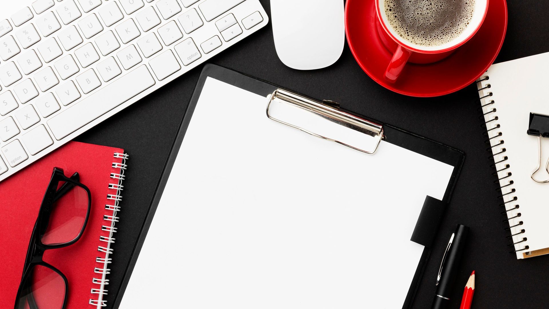 Black desk with keyboard, mouse, clipboard, coffee, red notebook, and glasses.