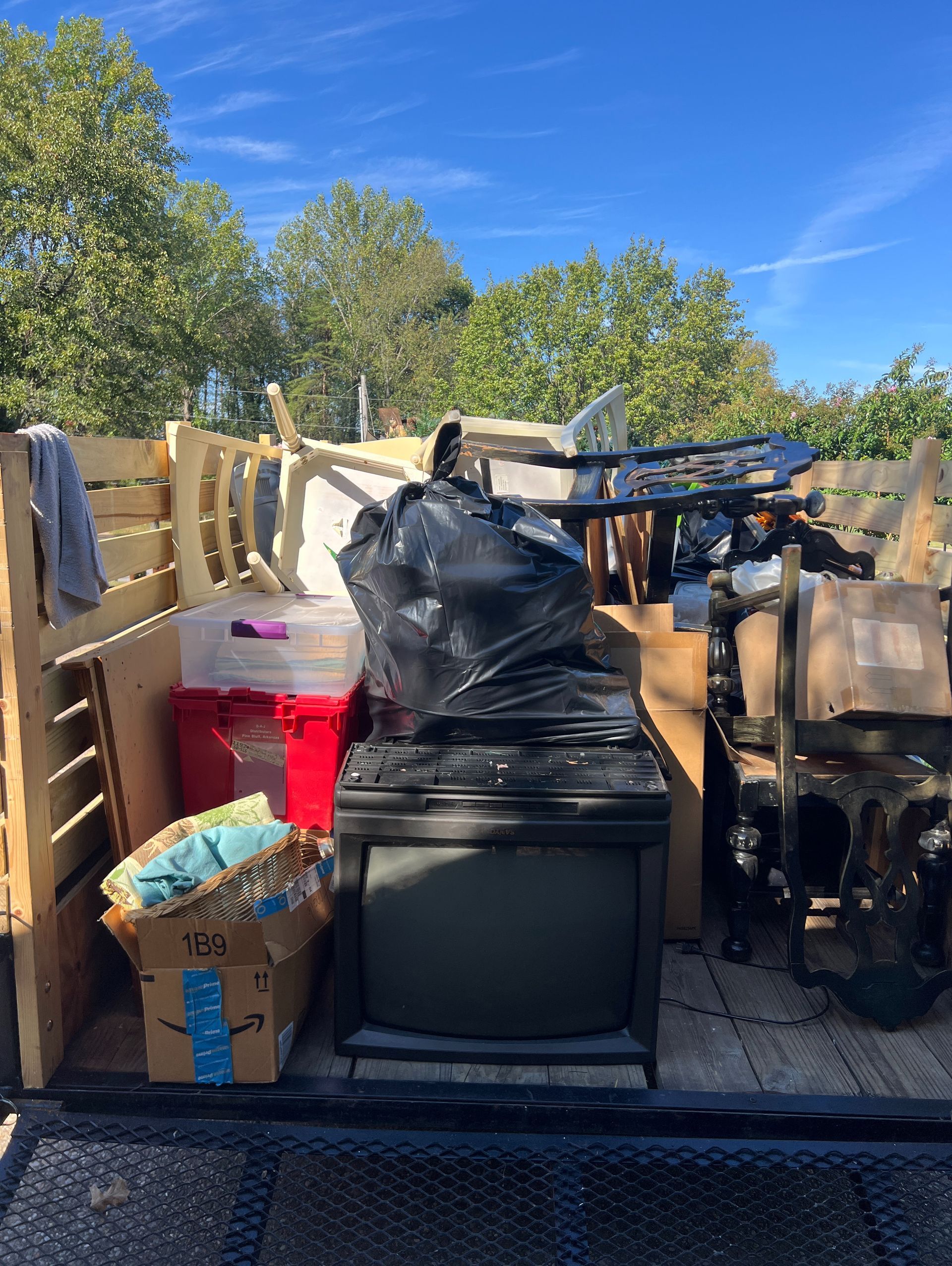Outdoor pile of discarded furniture, boxes, and trash piled along a fence under a bright blue sky.