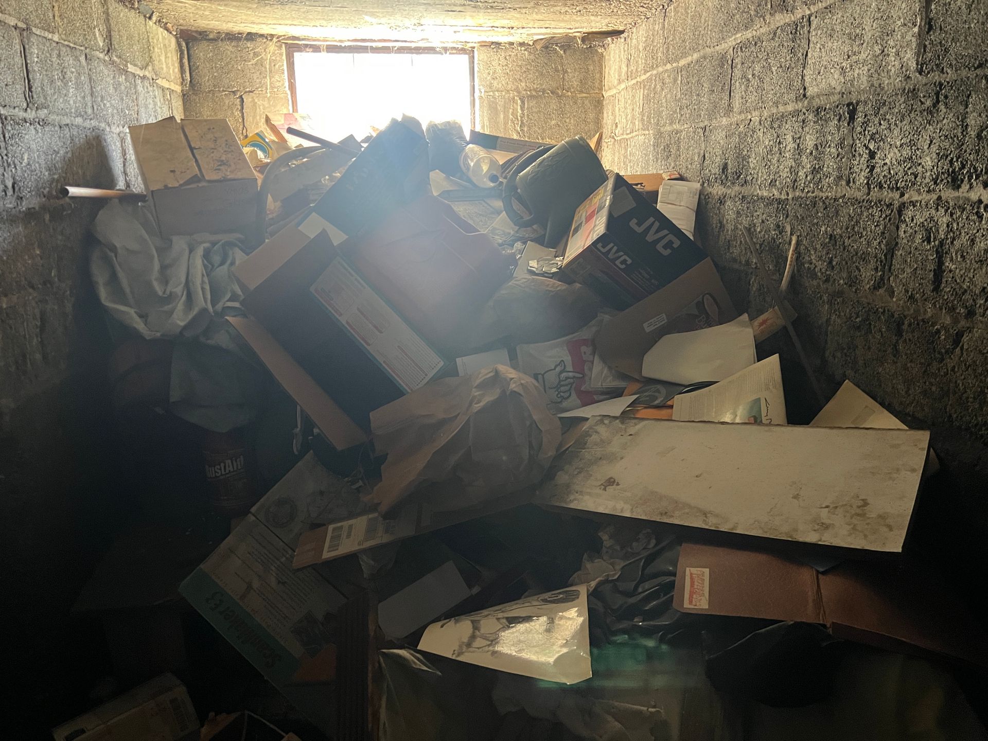 Dim cluttered basement with a large pile of cardboard, wood, and debris against concrete block walls.
