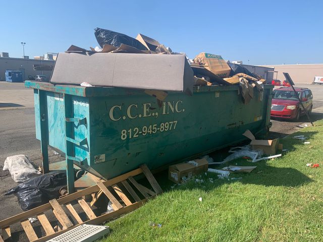 Green construction dumpster overflowing with debris beside a red car on a grassy roadside.