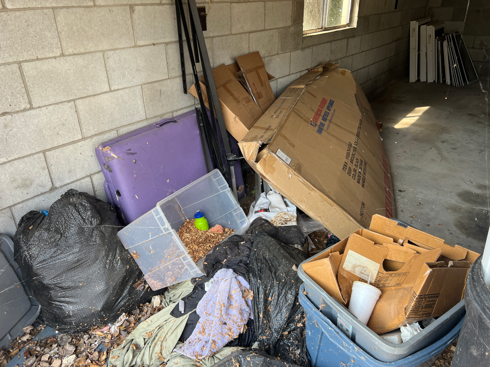 Messy garage corner with overflowing bins, cardboard, and scattered trash against cinderblock walls
