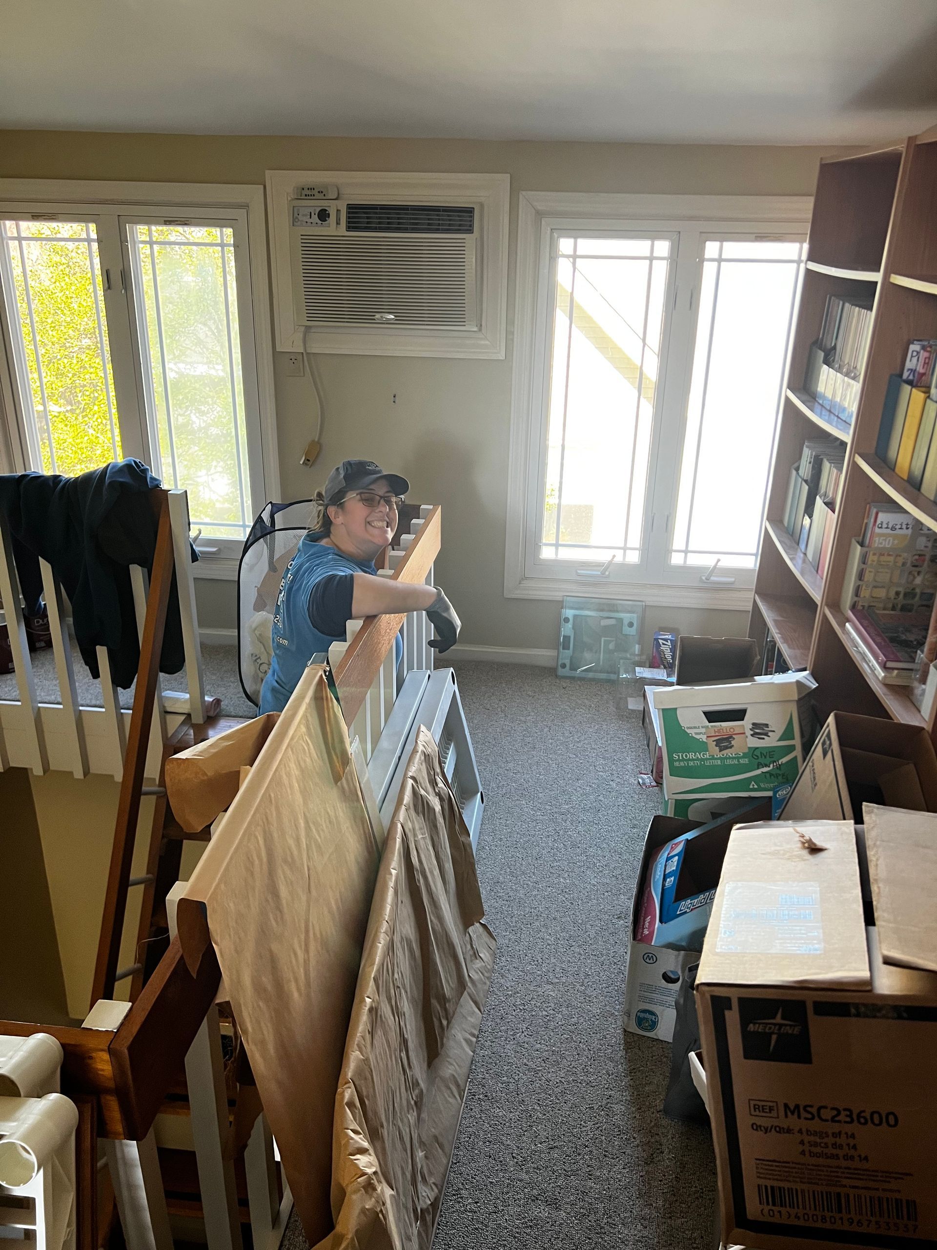 Person in a cluttered room stacking or moving items near windows and shelves.