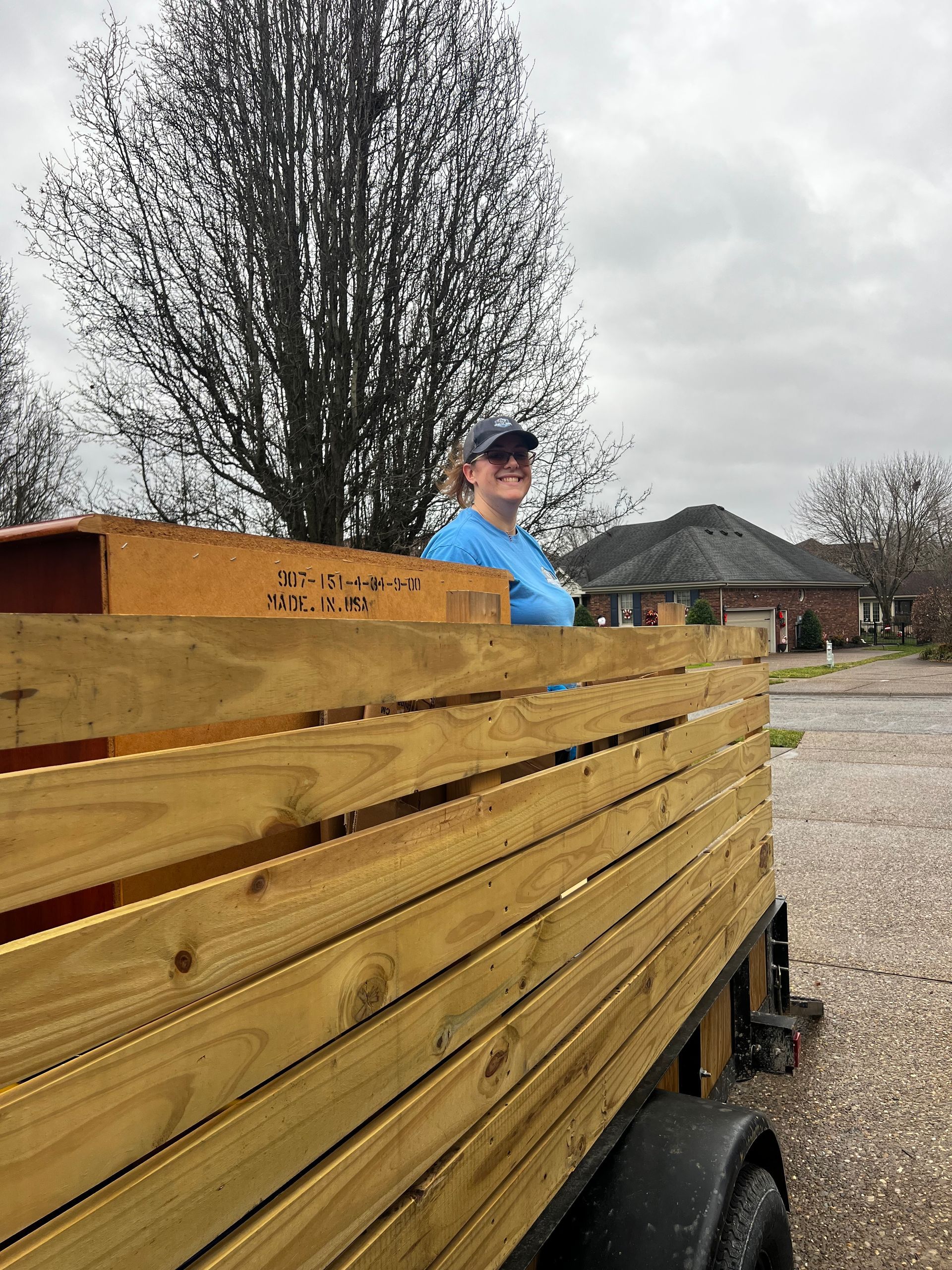 Person in a blue shirt sitting on a wooden trailer loaded with stacked lumber outdoors on a gravel lot.