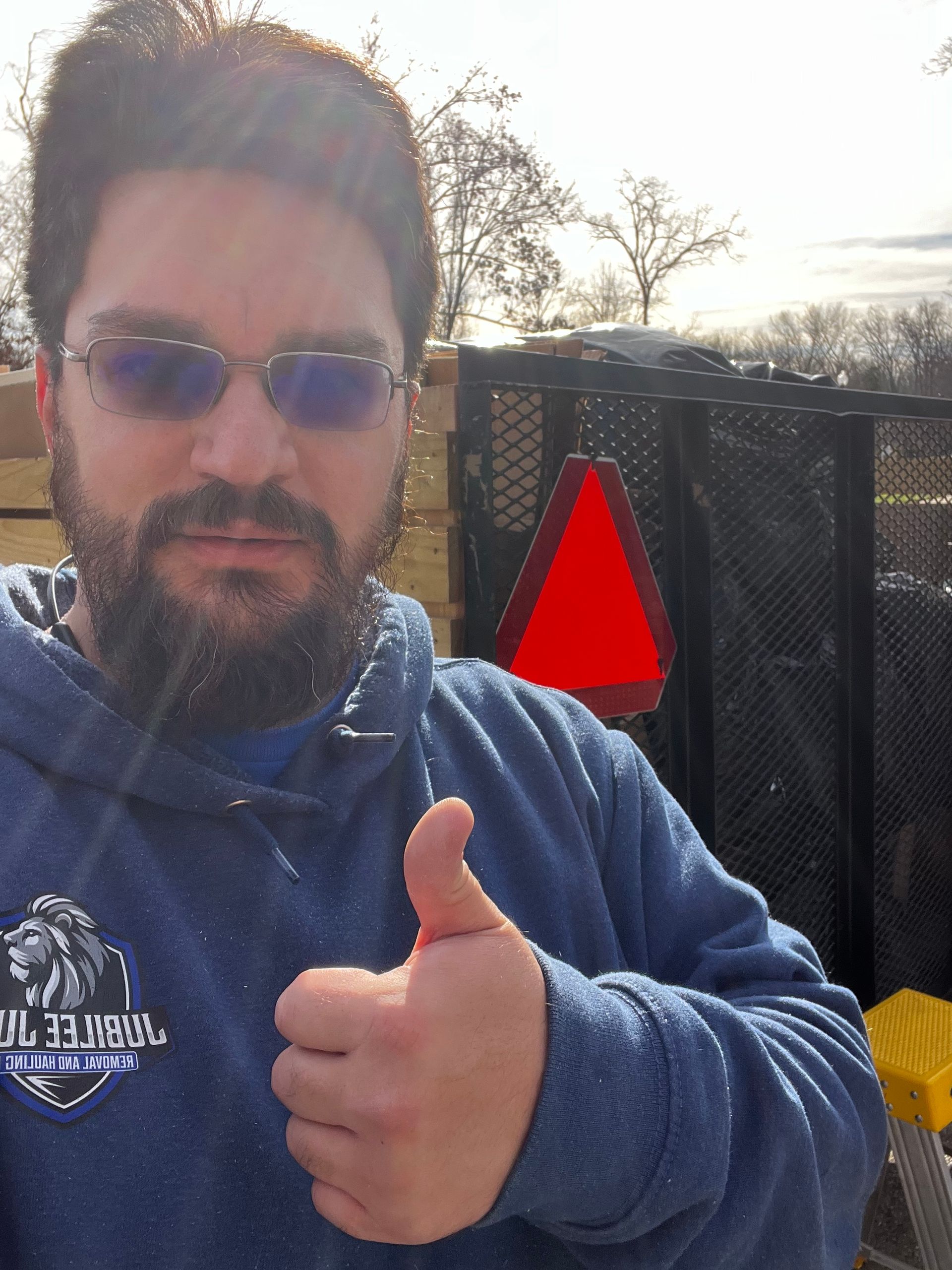 Man in blue hoodie and sunglasses giving a thumbs-up outdoors beside a black fence and red sign