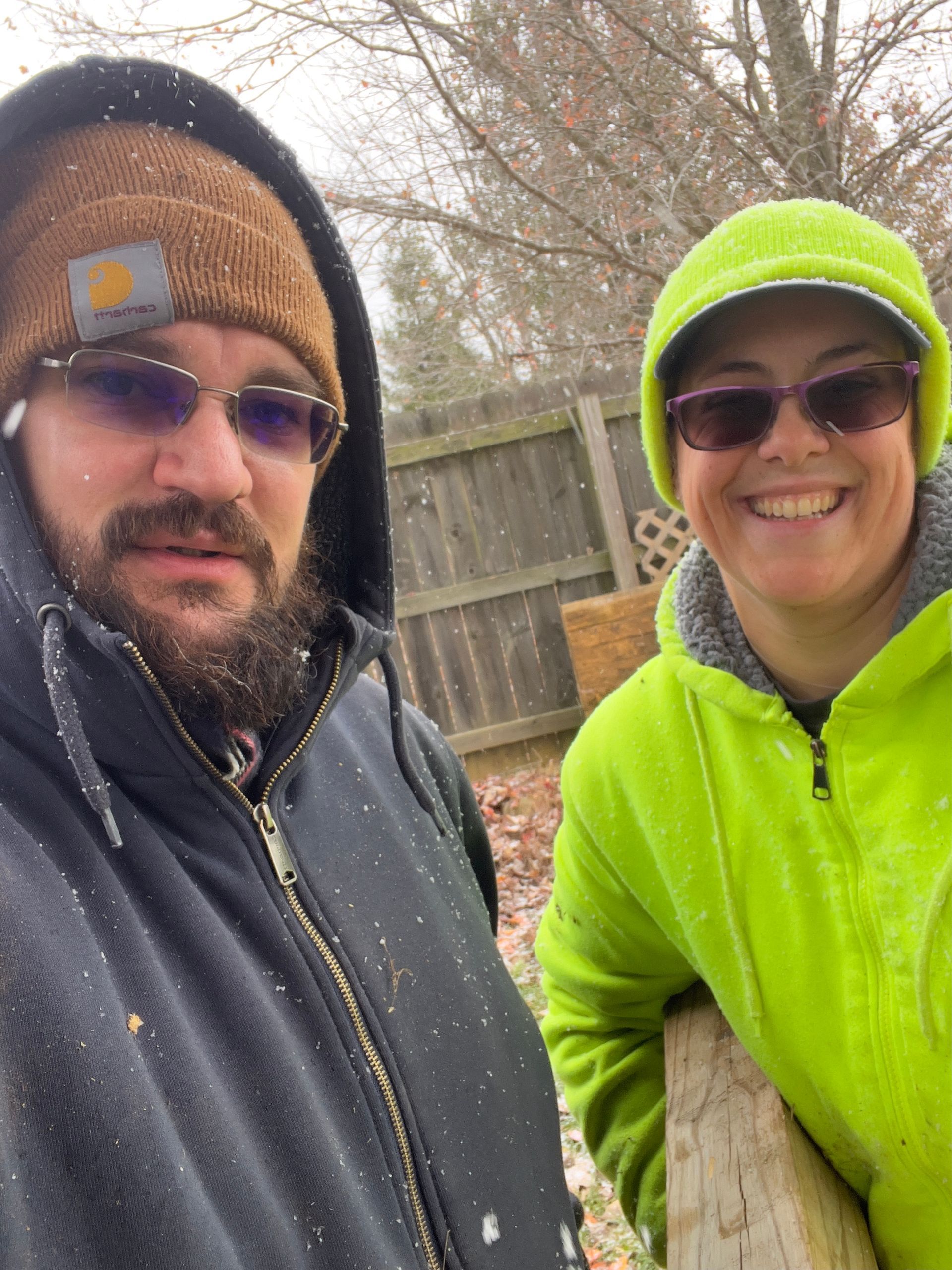 Two people in winter hats and jackets smile outdoors near a wooden fence.