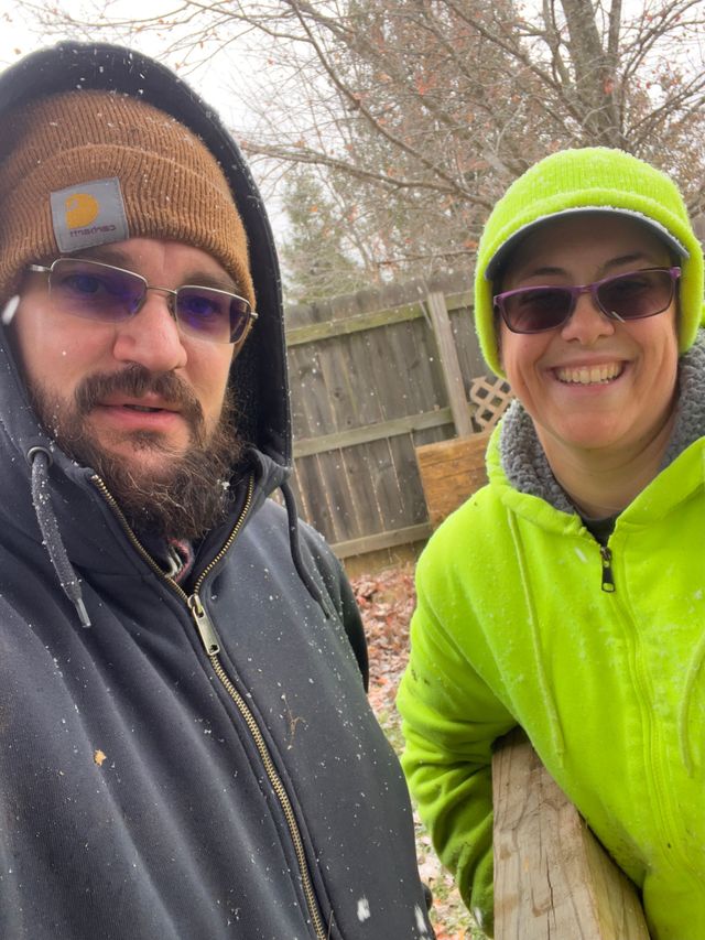 Two people in winter hats and jackets smile outdoors near a wooden fence.