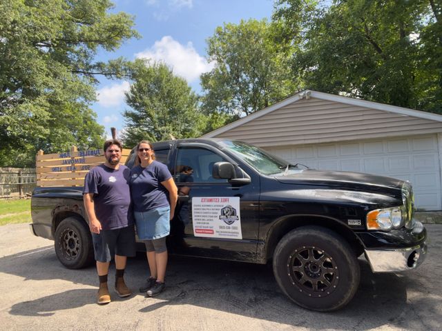 Two people standing beside a black pickup truck with a sign on the door in a sunny outdoor setting