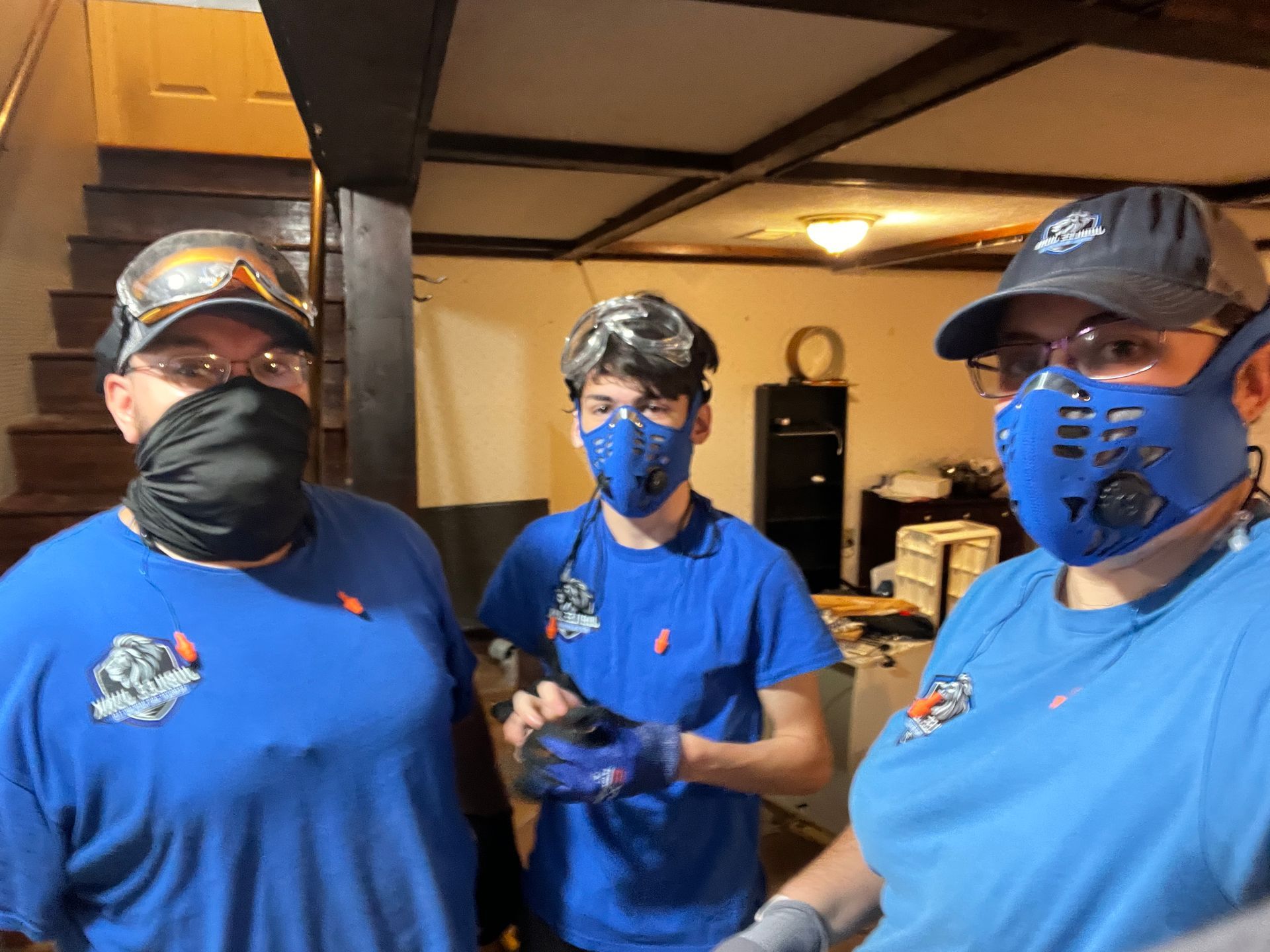 Three people in blue shirts and face masks standing indoors in a basement-like room.