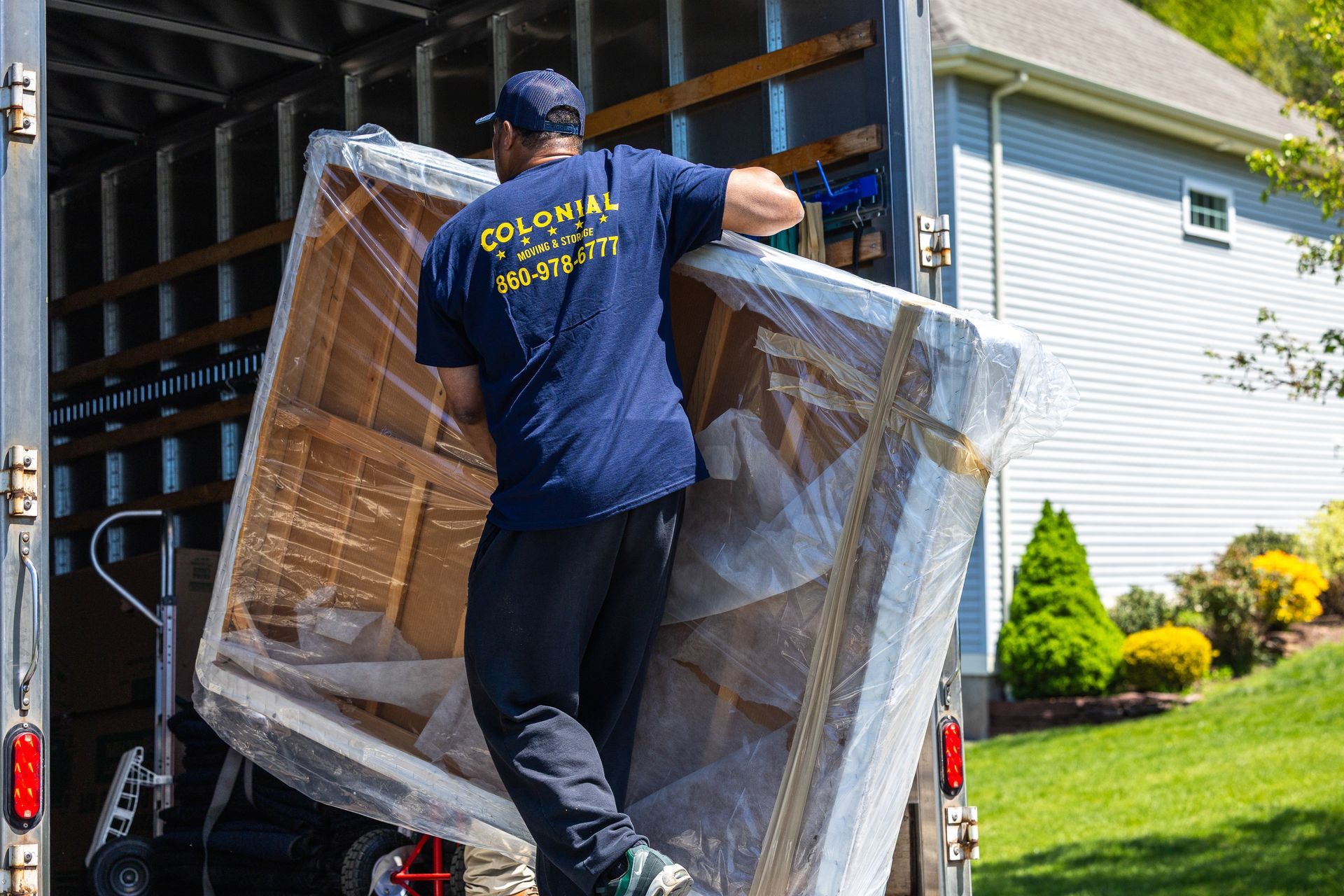 Mover carrying a wrapped mattress into a truck beside a house