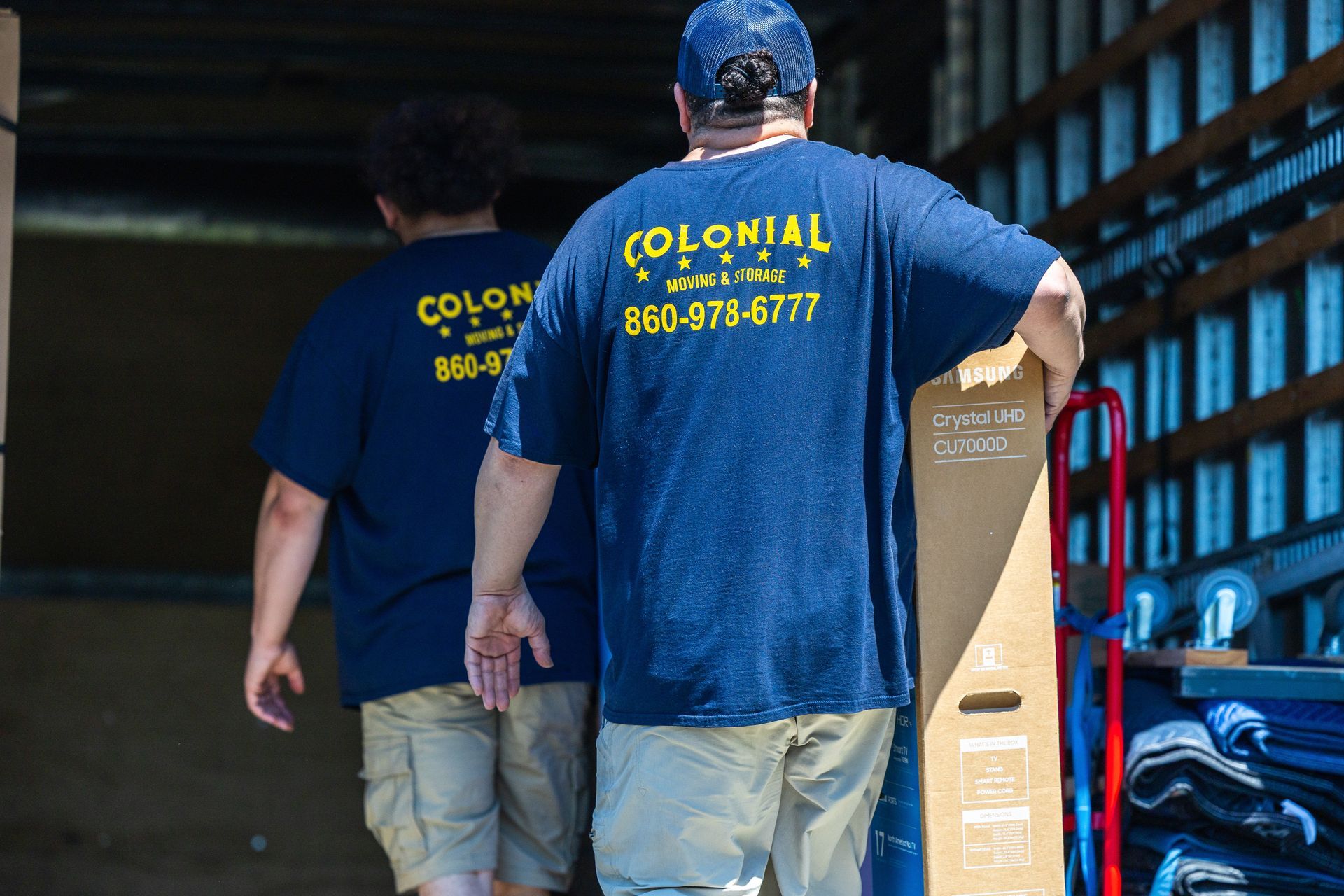 Two workers in blue shirts move items in a storage area, one carrying a large box.