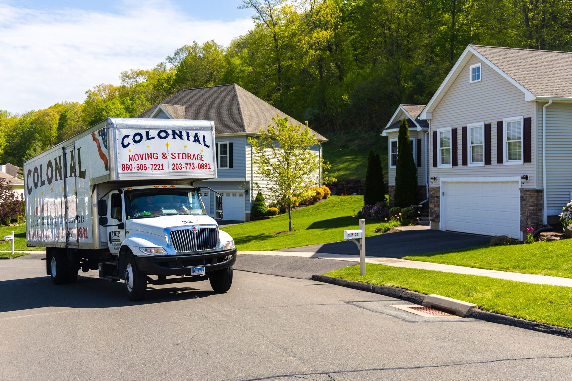Moving truck parked on a suburban street beside a white house and green lawn.