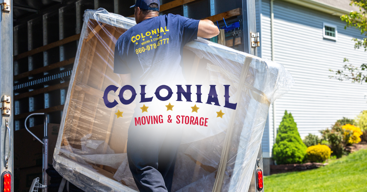 Mover carrying a large wrapped item through a Colonial Moving & Storage truck, outdoors by a house