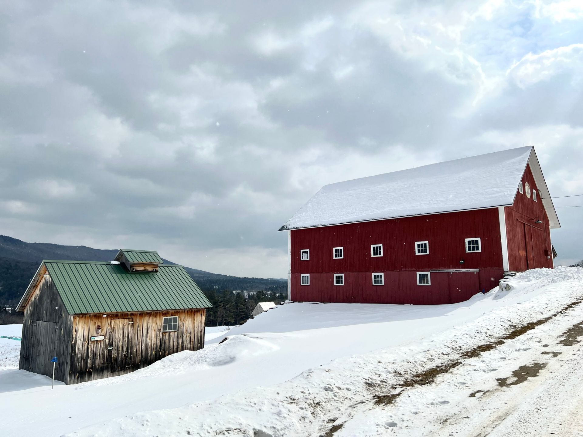 Red barn and small weathered shed on a snowy hillside under cloudy skies